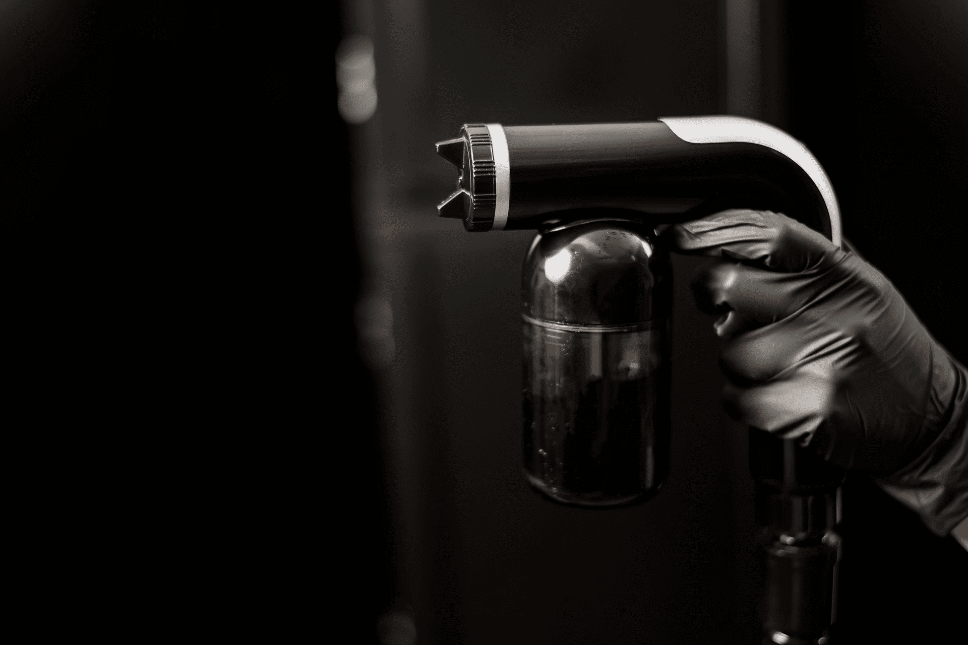 Hand in black glove holding a spray gun with a black and silver design against a dark background.